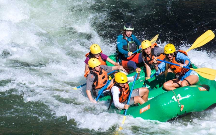 a group of students wearing helmets and life jackets paddle a green raft through whitewater